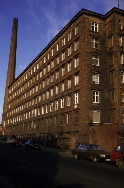 A tall brick industrial building with numerous windows on multiple floors stands beside a very tall brick smokestack under a blue sky.