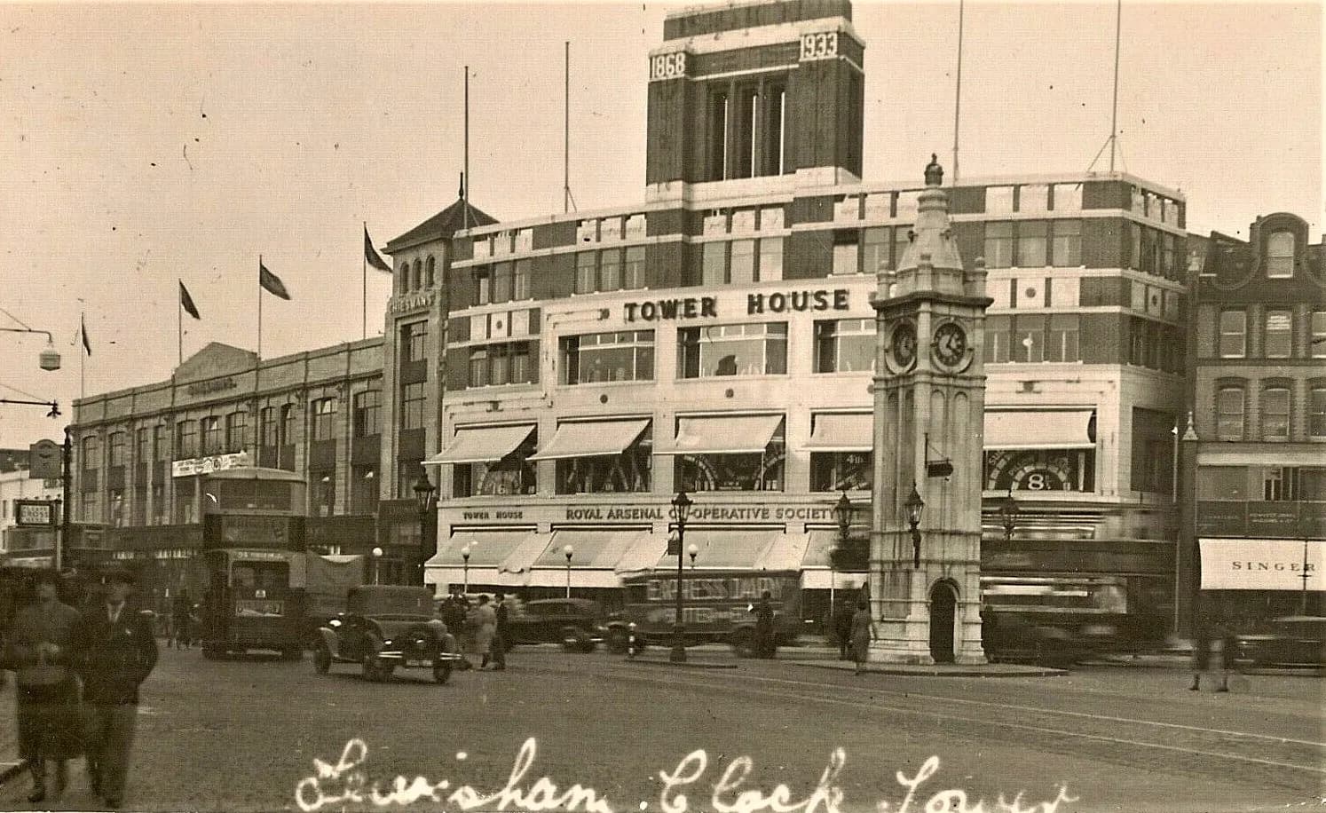 A bustling street scene in Lewisham, London, with the prominent clock tower and the 'Tower House' building of the Royal Arsenal Co-operative Society, along with other shops and vintage vehicles.