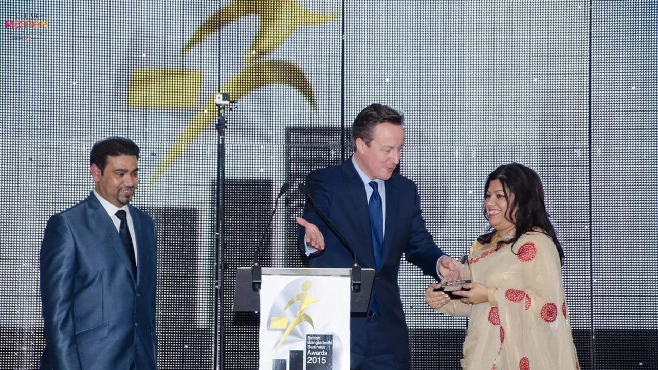 A man in a suit standing at a podium handing an award to a smiling woman in a sari, with another man in a suit standing to the left.