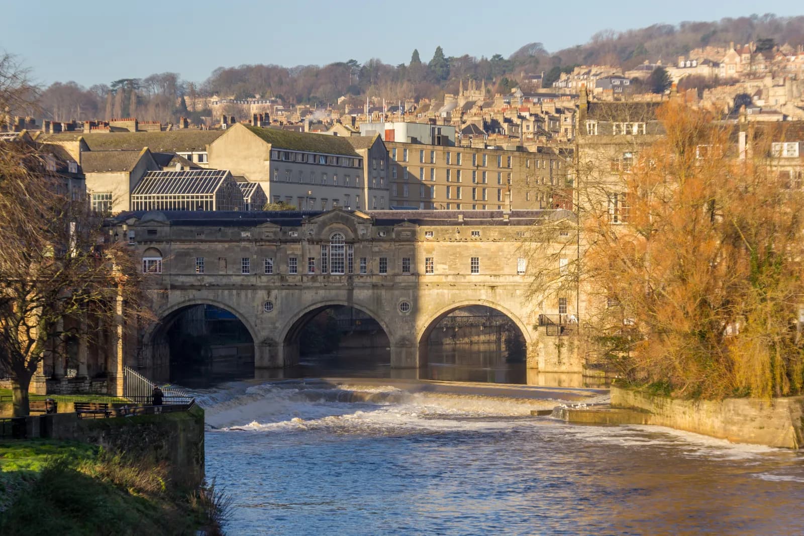 Pulteney Bridge spans a river of flowing water with residences built into it and hills covered in buildings and trees behind it.