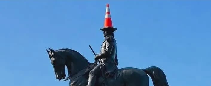 A black equestrian statue of a historical figure, possibly George Washington, with an orange traffic cone placed on its head, set against a clear blue sky.