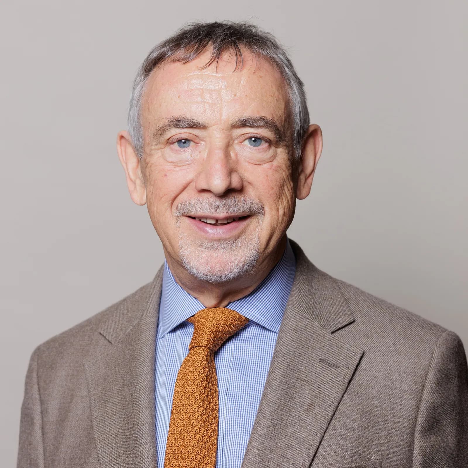 A smiling man with short grey hair and a goatee beard wears a light brown jacket over a blue and white striped shirt with an orange knit tie.