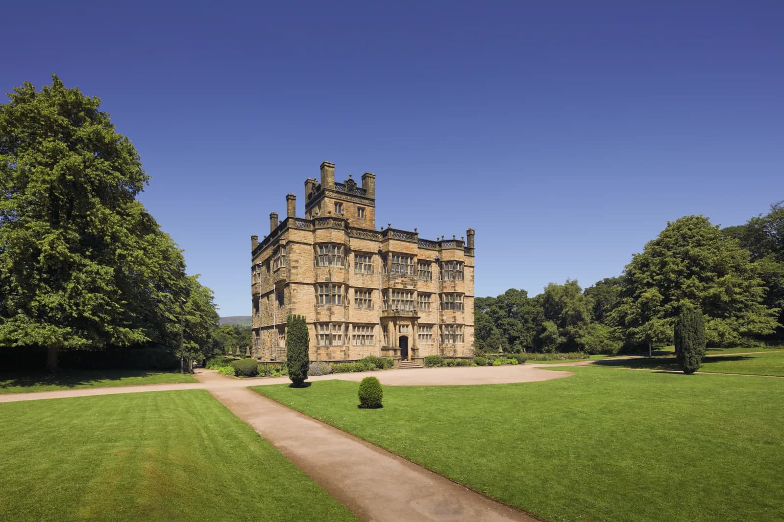 A grand, multi-story stone manor with intricate window detailing and turrets stands majestically amidst expansive green lawns and lush trees under a clear blue sky.