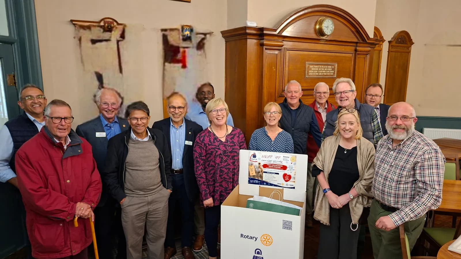 A group of smiling men and women of various ages stand around a white box with a "Rotary" logo and a sign for "Wrap Up" with a heart icon.