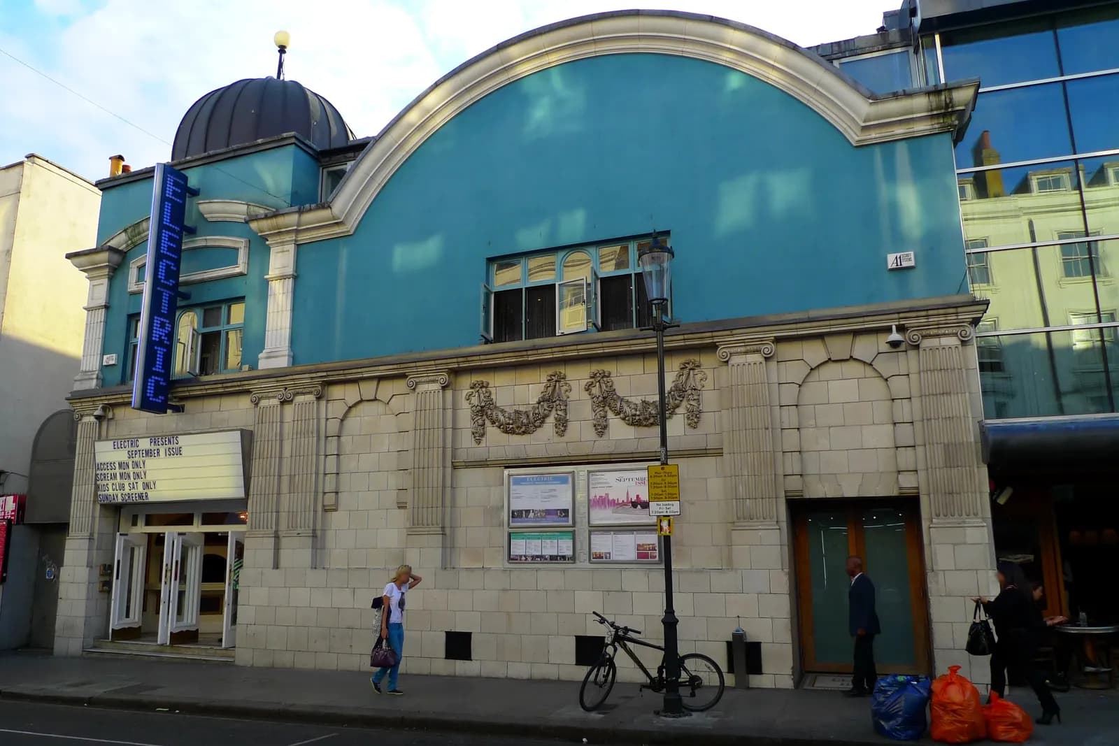 A light blue building with a domed roof and a neon sign for "ELECTRIC" on the left, an entrance with a movie marquee advertising "ELECTRIC PRESENTS SEPTEMBER ISSUE," and people and a bicycle on the sidewalk in front.