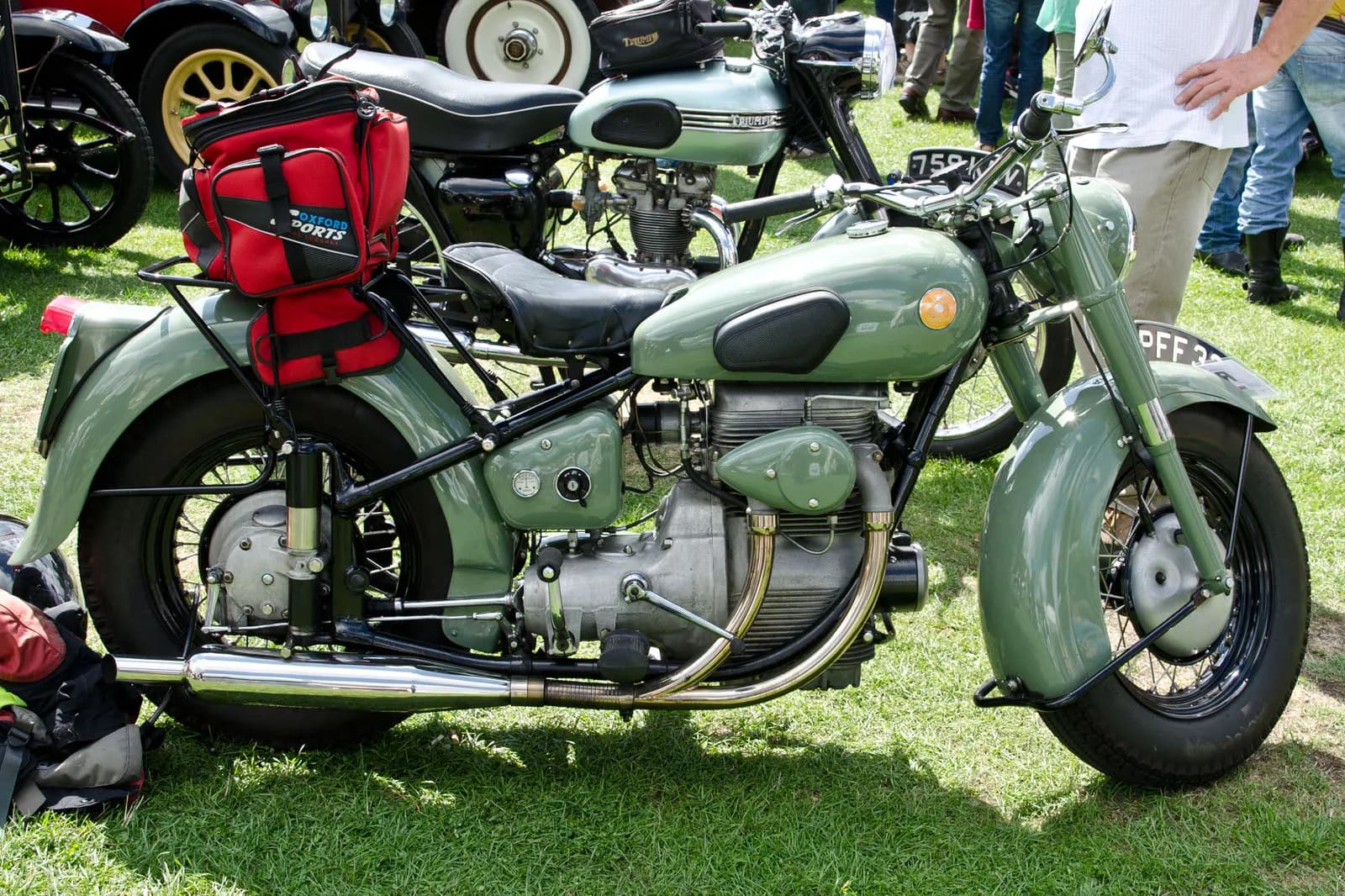 A light green vintage motorcycle with a red bag on the back is parked on a grassy field, with other motorcycles and people visible in the background.