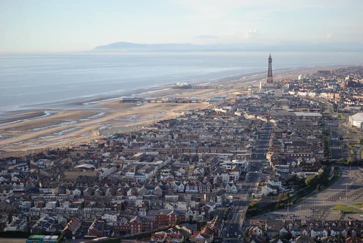 An aerial view of a coastal city, showing a dense collection of buildings, a sandy beach, and a pier extending into the calm ocean under a clear sky.