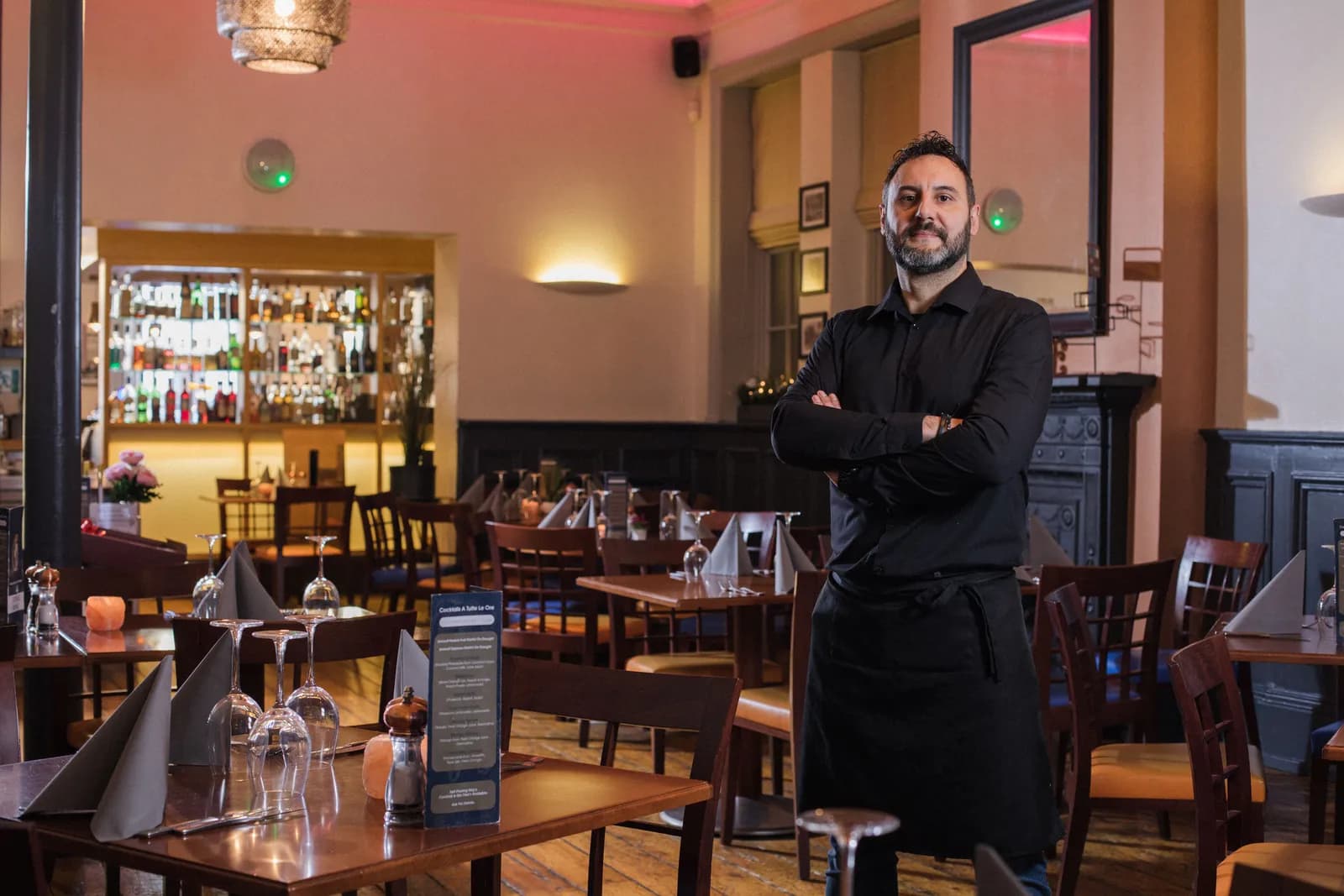 A man with a beard and dark shirt stands with his arms crossed in a restaurant, with dining tables and a bar in the background.