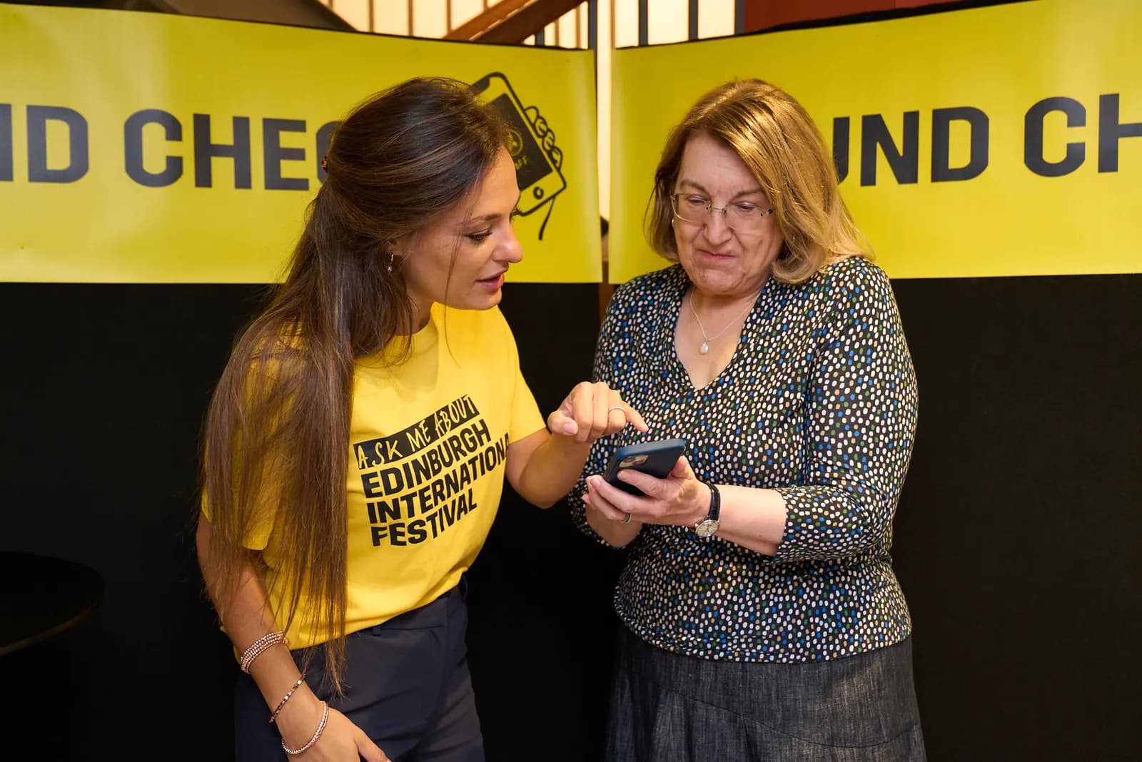 Two women, one in a yellow t-shirt and the other in a patterned blouse, look at and interact with a smartphone held by the woman in the patterned blouse, against a yellow and black backdrop.