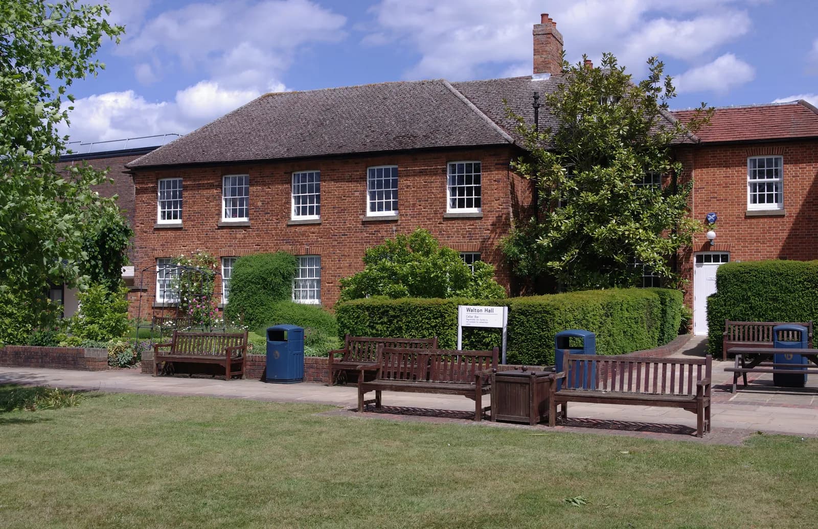 A red brick building with many windows sits behind a manicured green hedge and a small lawn with wooden benches.