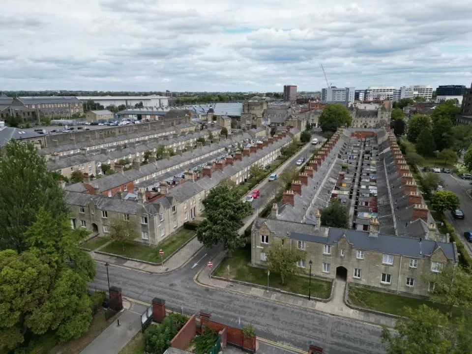 An aerial view of residential streets with rows of terraced houses, surrounded by trees and a wider cityscape in the distance under a cloudy sky.