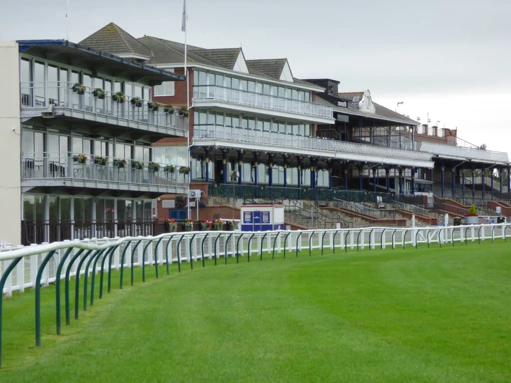 A large building with multiple levels of balconies and windows, overlooking a bright green grassy field with a white fence.
