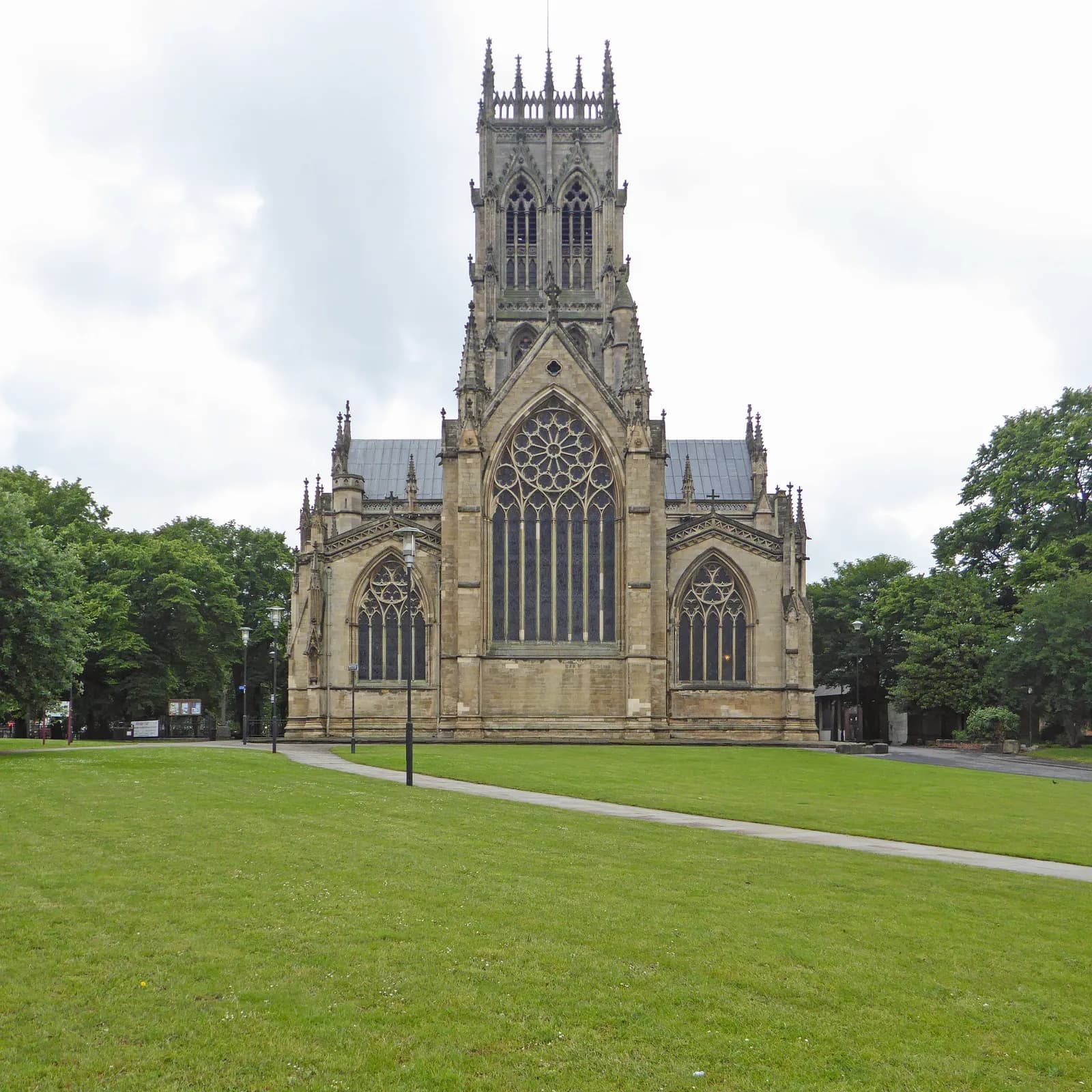 A grand Gothic cathedral with a tall, ornate bell tower and large stained-glass windows stands majestically behind a lush green lawn and walking path under a cloudy sky.