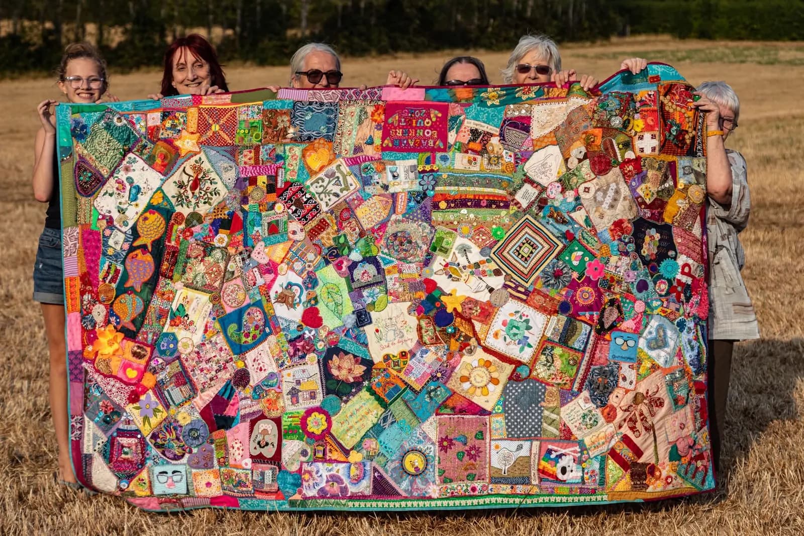 Six people holding up a large, vibrantly colored, patchwork quilt featuring various embroidered designs and fabric textures in an outdoor field.