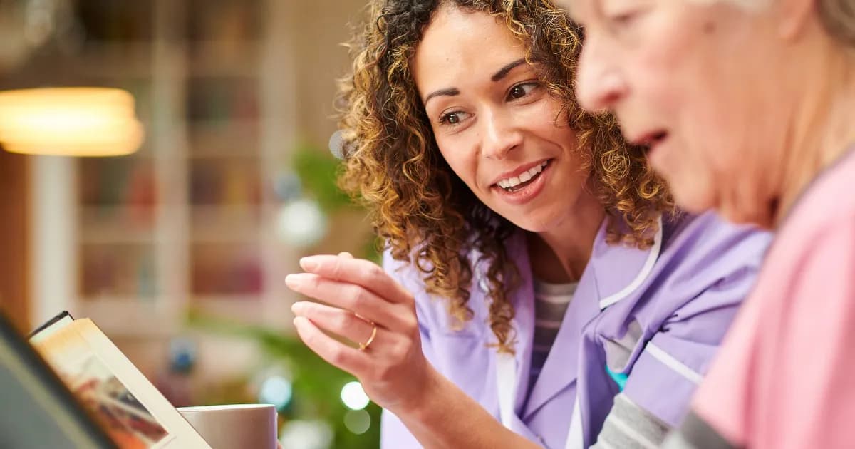 A smiling young caregiver with curly hair in a purple uniform gestures as she looks at an elderly person who is partially visible on the right, suggesting conversation or shared activity.
