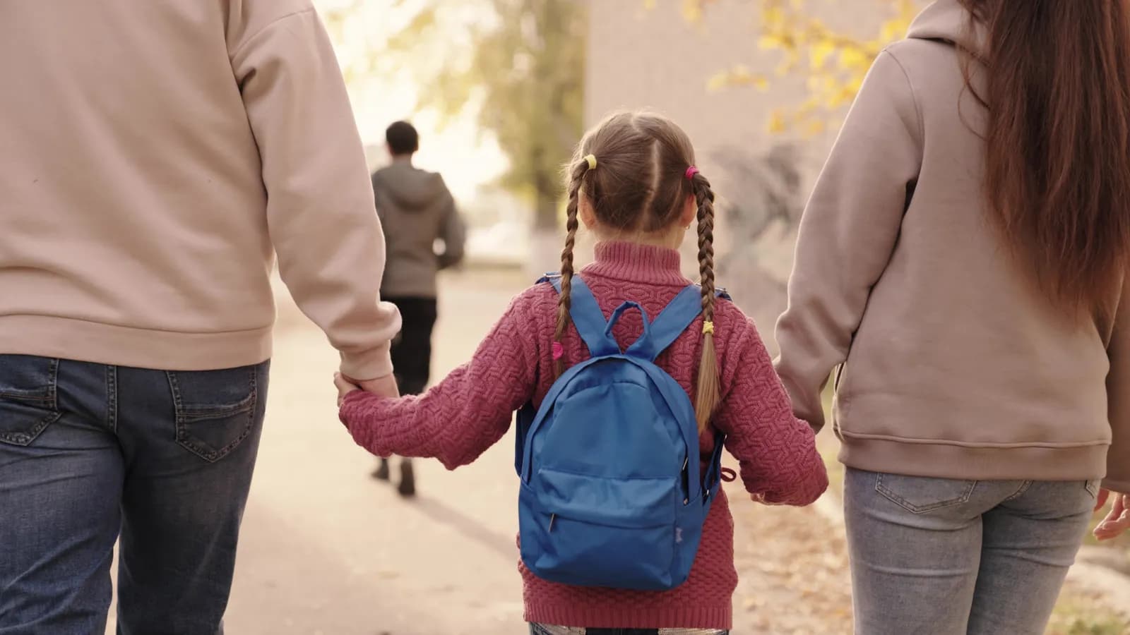 A young girl with pigtails and a blue backpack holds hands with two adults as they walk away, with another person visible in the background.