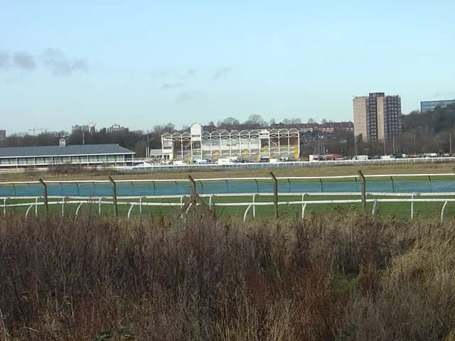 A wide shot shows a racetrack with a blue track and white railing in the foreground, with buildings, including a grandstand and apartment blocks, in the background.