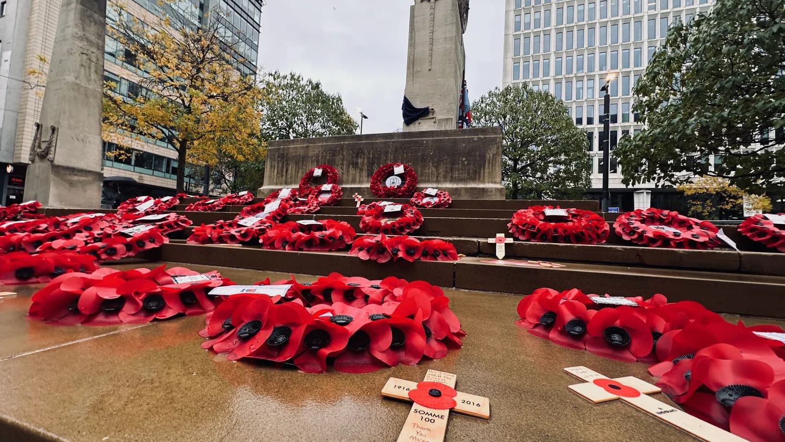 Red poppy wreaths and wooden crosses commemorating soldiers are arranged on the steps of a stone war memorial in a city.