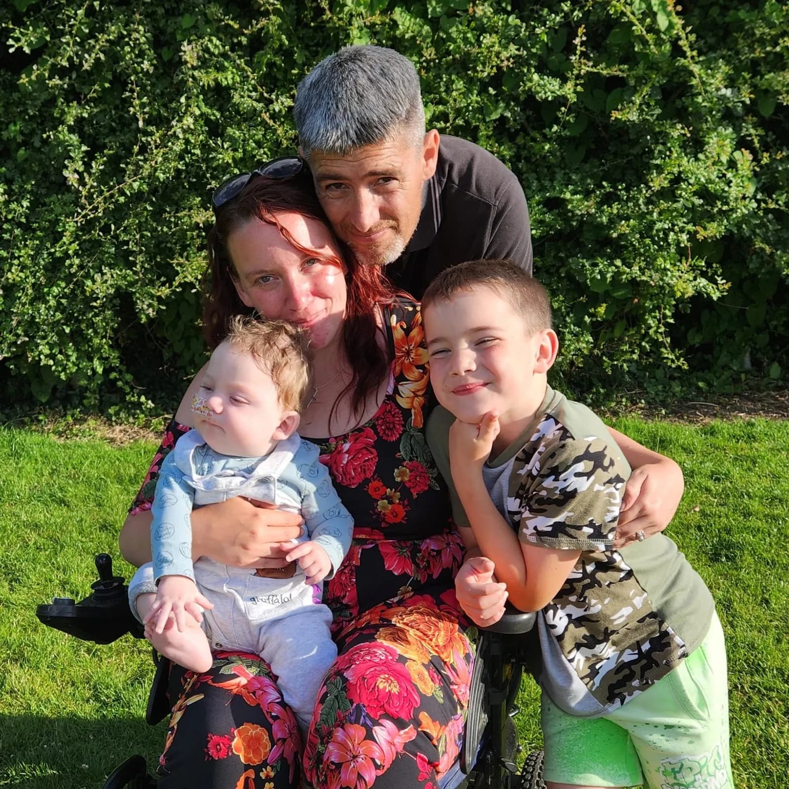 A family of four, including a mother in a wheelchair holding a baby, a father, and an older boy, poses outdoors with a green hedge behind them.