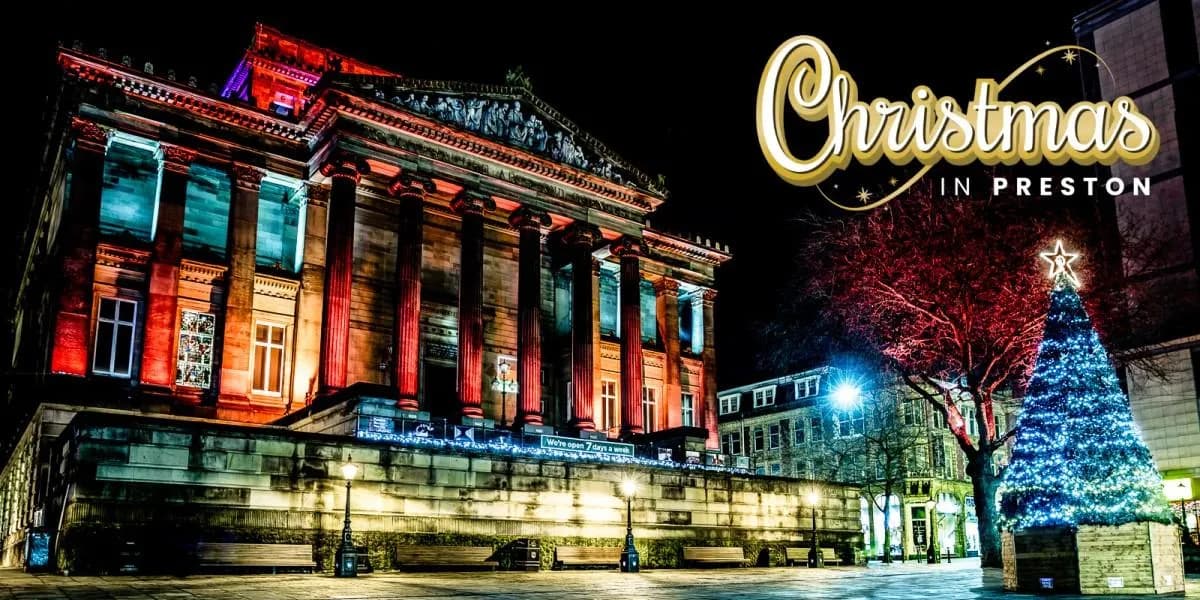 A classical building with tall pillars is illuminated with red and blue lights at night, next to a brightly lit blue Christmas tree and a tree with red lights, all under a dark sky with a "Christmas in Preston" graphic.