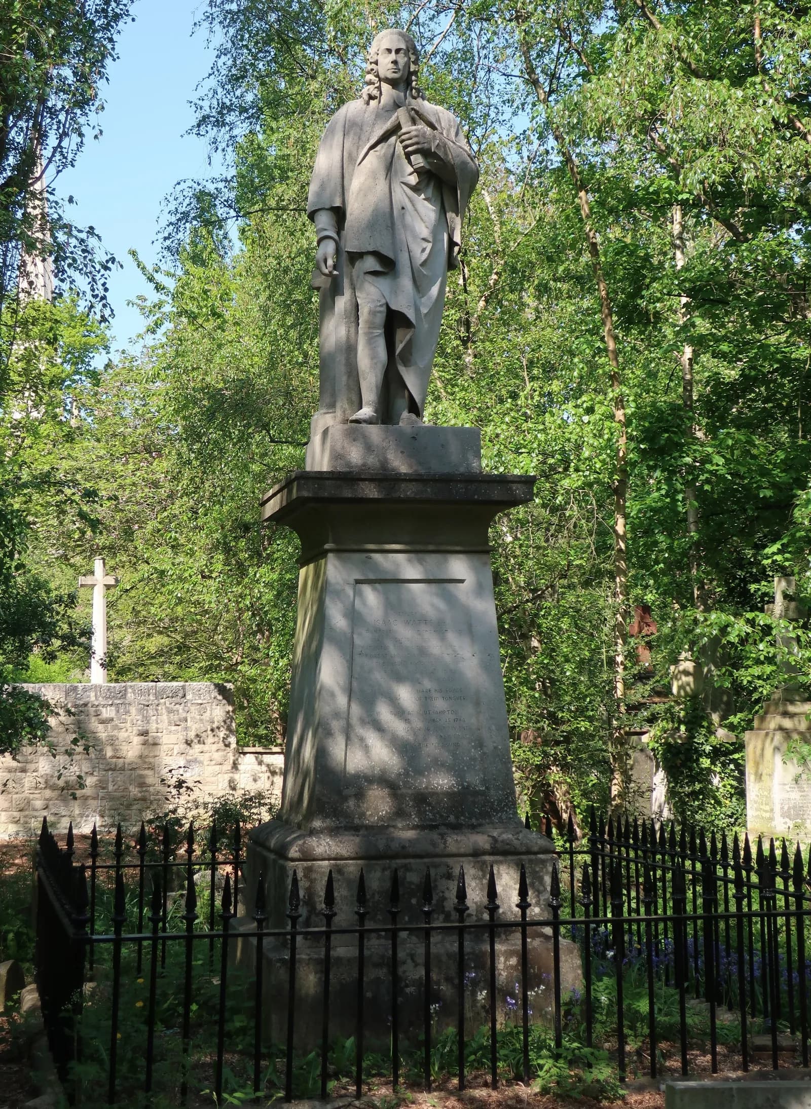 A tall stone statue of a man with long hair, wearing a loose robe and holding an object, stands on a pedestal surrounded by green trees and a black metal fence, with a stone wall and a white cross visible in the background to the left.