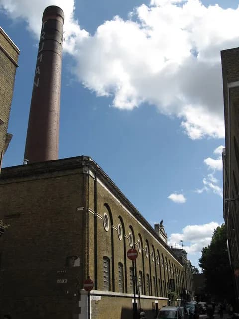 A tall brick chimney labeled 'TRUMAN' stands beside a long brick building with arched windows, under a bright blue sky with white clouds.