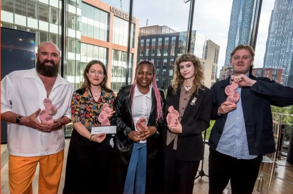 A group of five people, four women and one man, holding pink art awards inside a modern building with large glass windows overlooking a city.