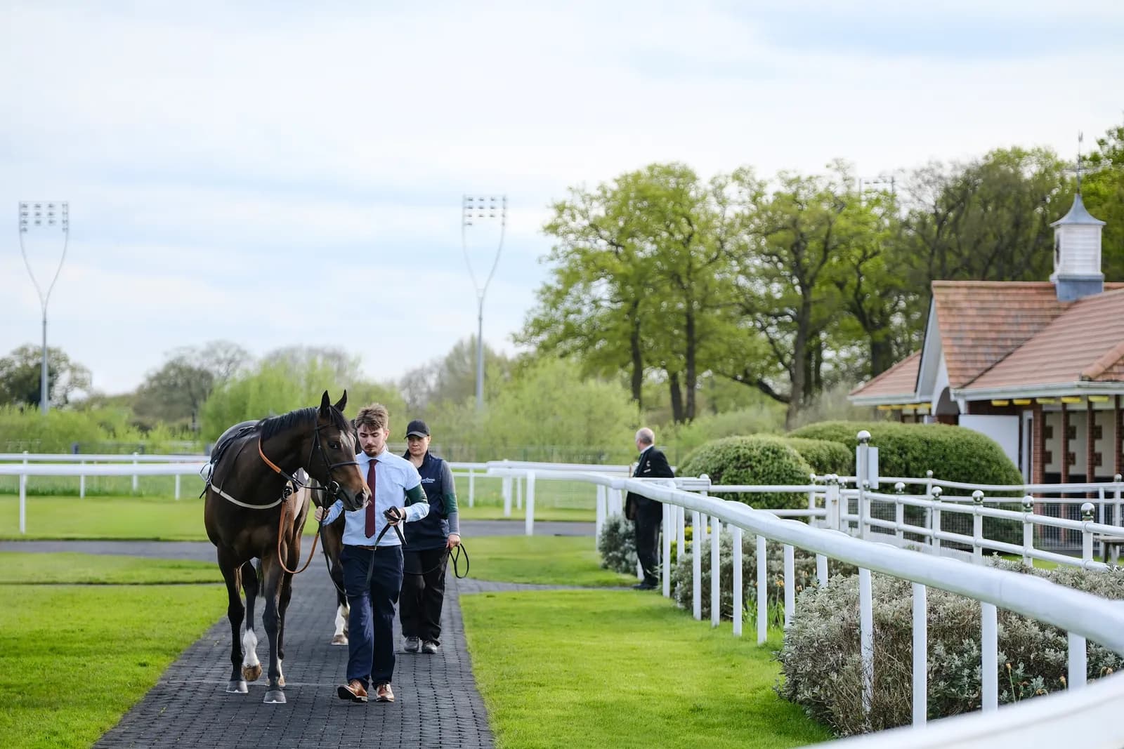 A horse and two people walk along a paved path next to a green race track, with a small building and trees in the background.