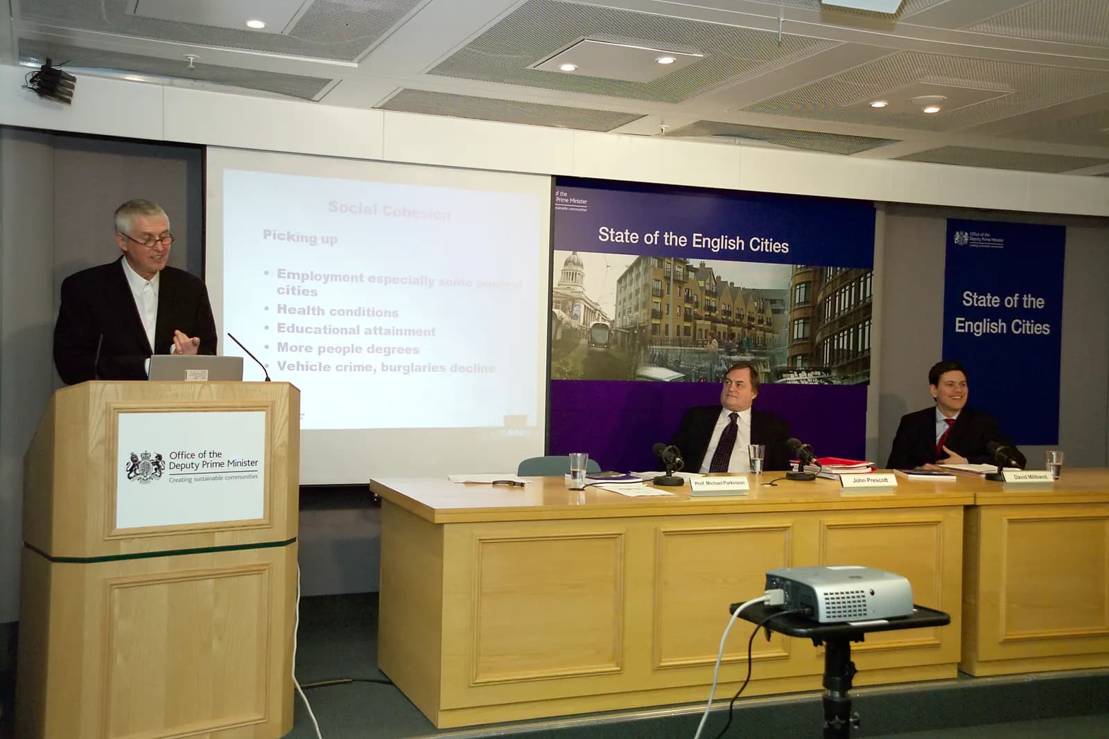 A man in a suit speaks at a podium with the 'Office of the Deputy Prime Minister' logo while two other men sit at a table in front of a banner that reads 'State of the English Cities'.