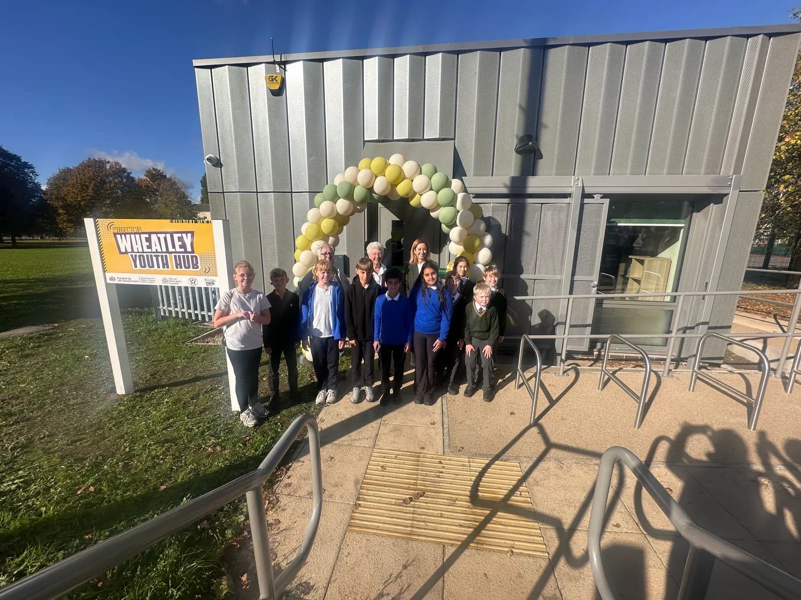 A group of children and adults stand in front of the Wheatley Youth Hub building, which has a balloon arch over its entrance and a sign to its left.