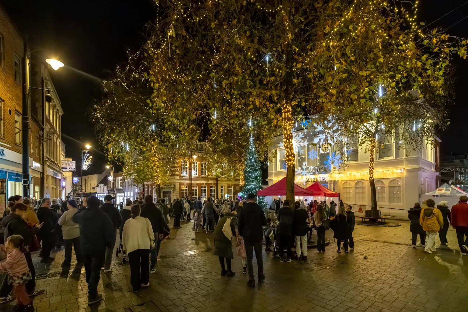 A nighttime street scene with a crowd of people, illuminated by festive lights strung across trees and buildings, with a red tent and a Christmas tree visible in the background.