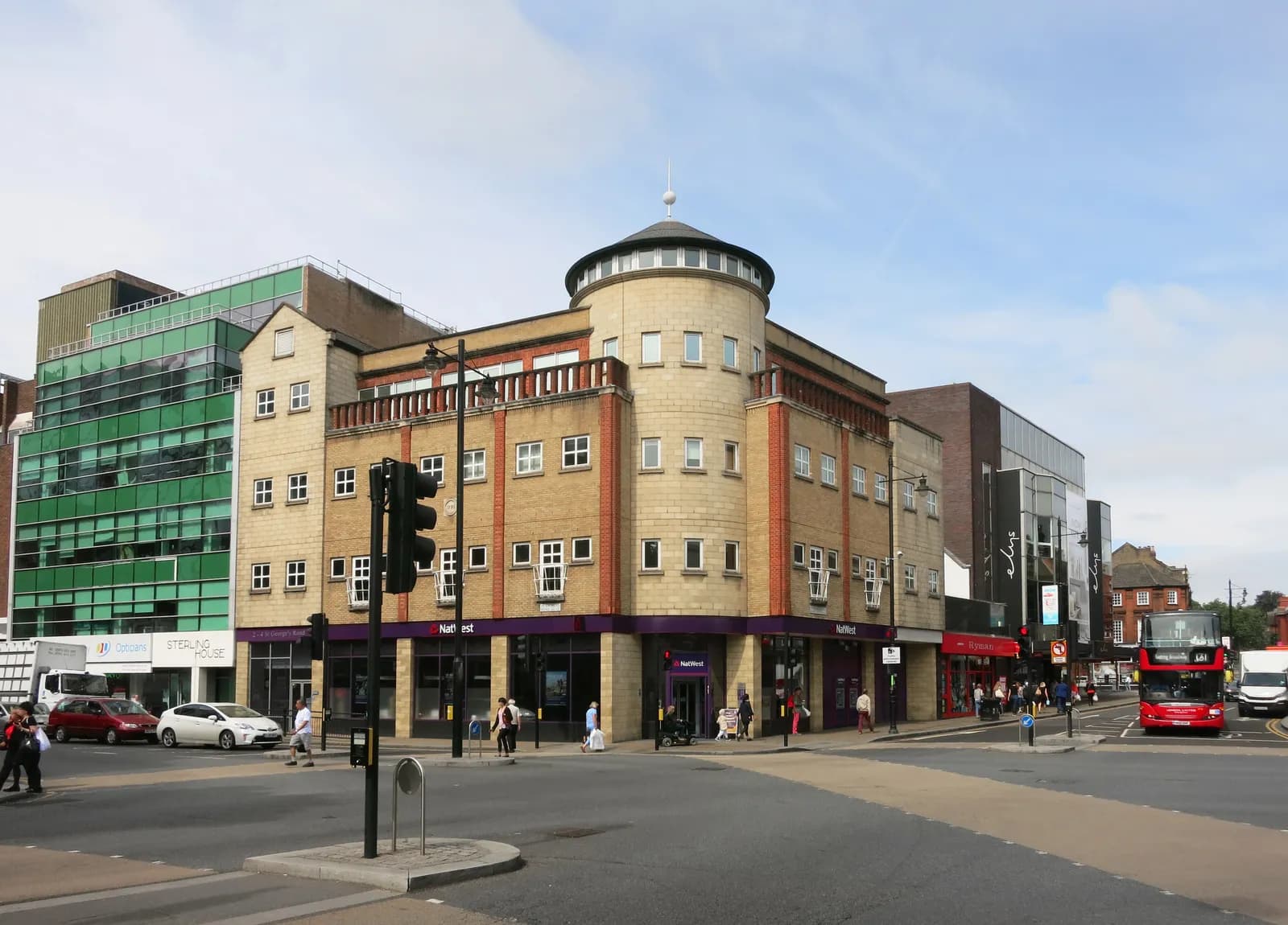 A bustling street scene with a light-colored commercial building at the intersection, featuring a rounded corner with a domed top, and a double-decker bus in the distance.