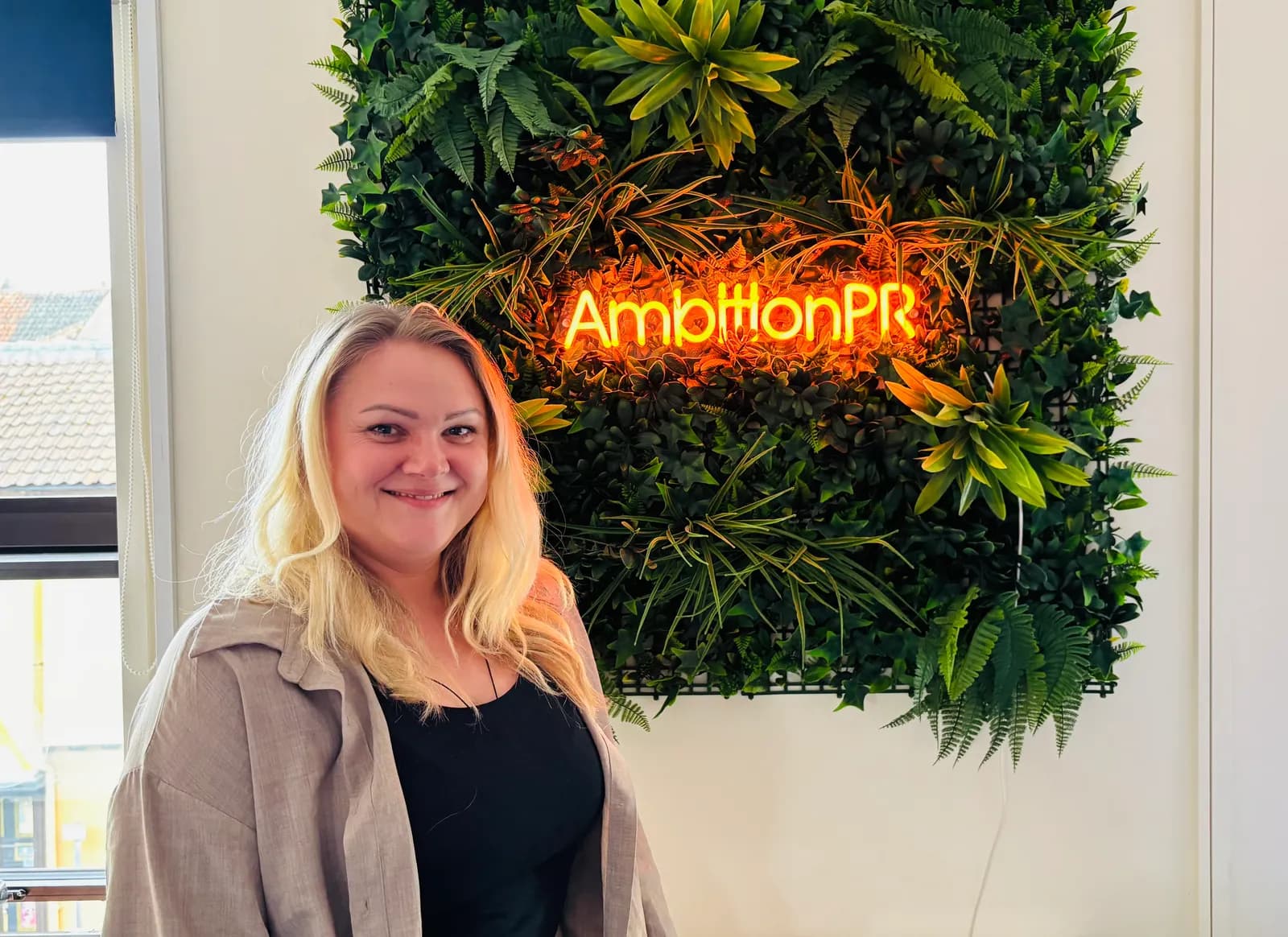 A smiling blonde woman stands in front of a green plant wall with an orange neon sign that reads "AmbitionPR."