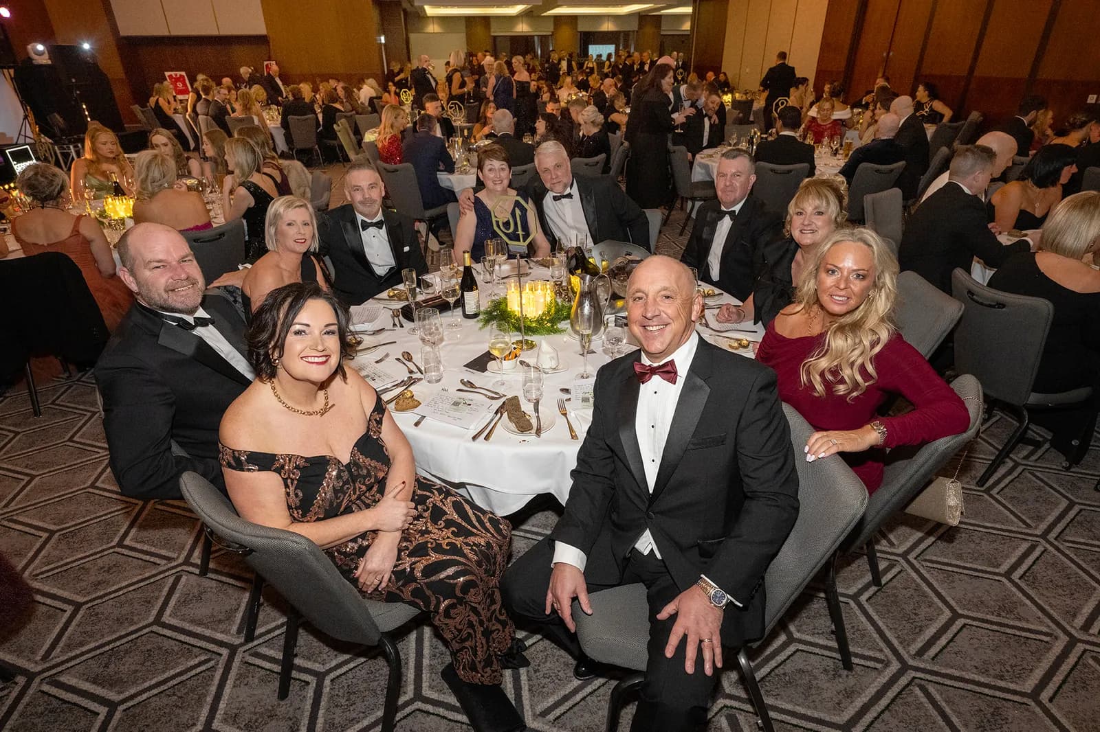 A group of formally dressed adults are seated at round tables in a large event hall, looking towards the camera.