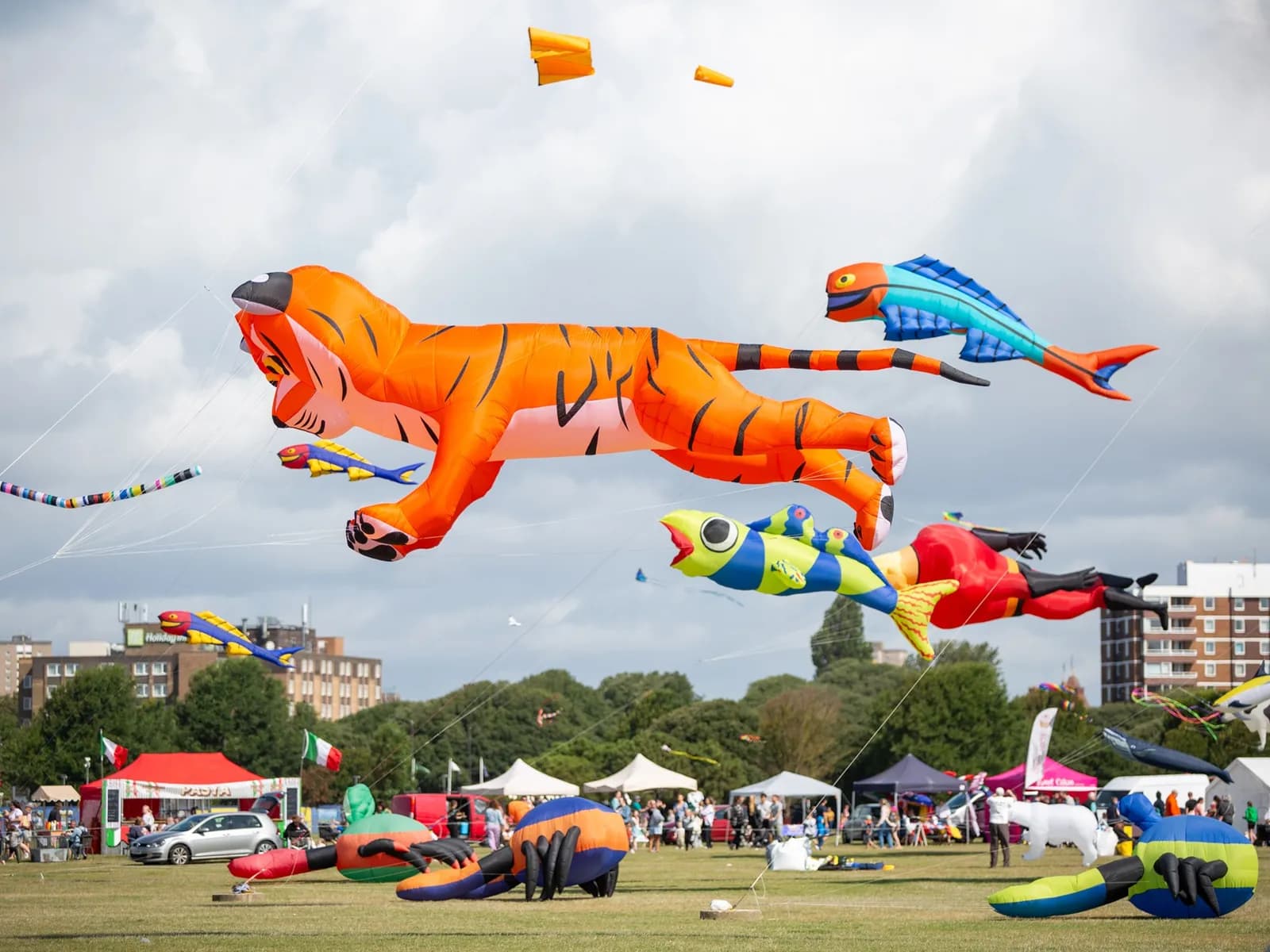 A large inflatable tiger kite flies in the sky over a grassy field with other smaller kites and tents in the background.