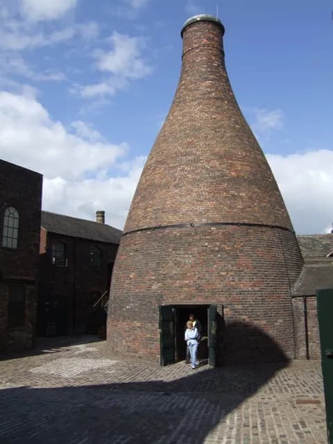 A woman stands in the doorway of a large, bottle-shaped brick pottery kiln, with another person visible behind her, in a paved courtyard under a partly cloudy sky.