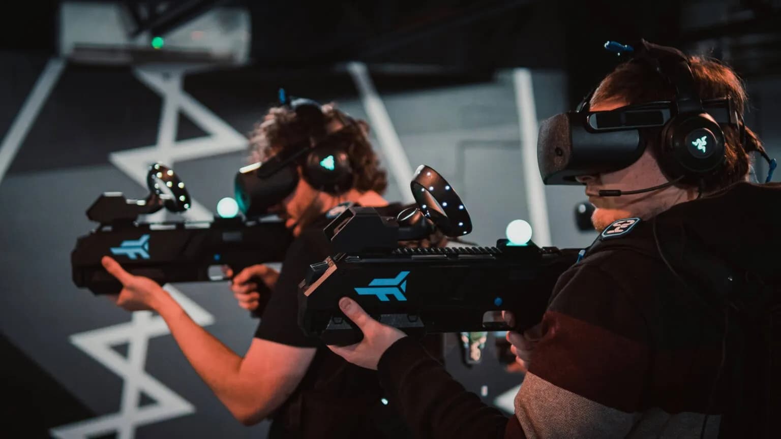 Two men wearing VR headsets and holding large virtual reality guns in an arcade.