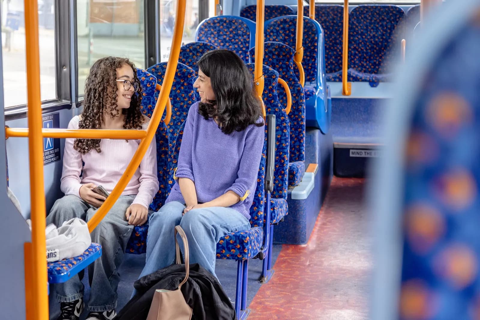 Two women, one with curly hair and glasses, sit facing each other on a public bus.