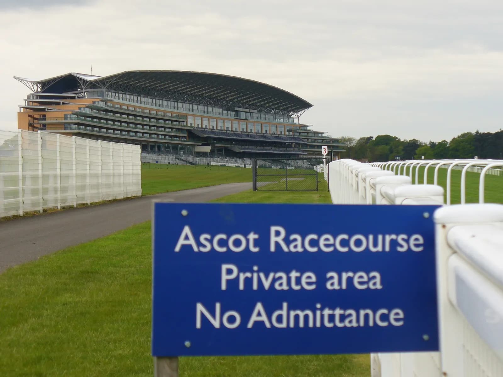 A blue sign reading "Ascot Racecourse Private area No Admittance" in the foreground with the grandstand building of Ascot Racecourse in the background under a cloudy sky.