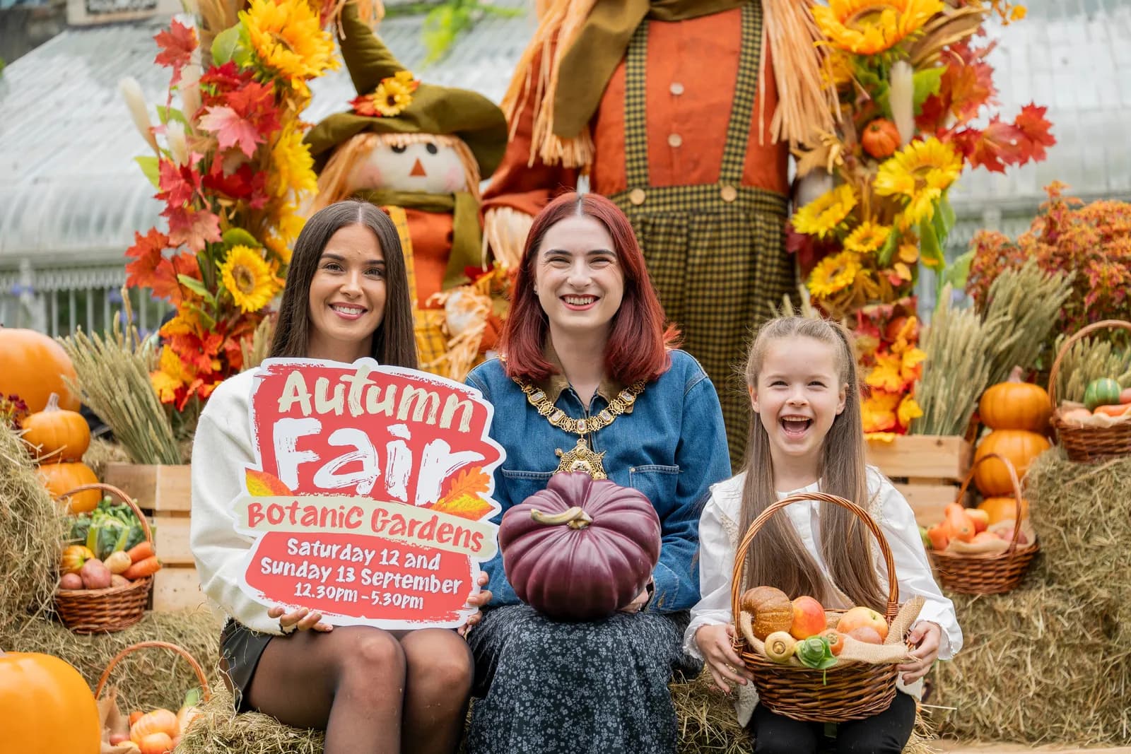Three smiling females, two adults and one child, sitting in front of autumn decorations including scarecrows, pumpkins, and hay bales. The adult on the left holds a sign for an 'Autumn Fair' at Botanic Gardens.