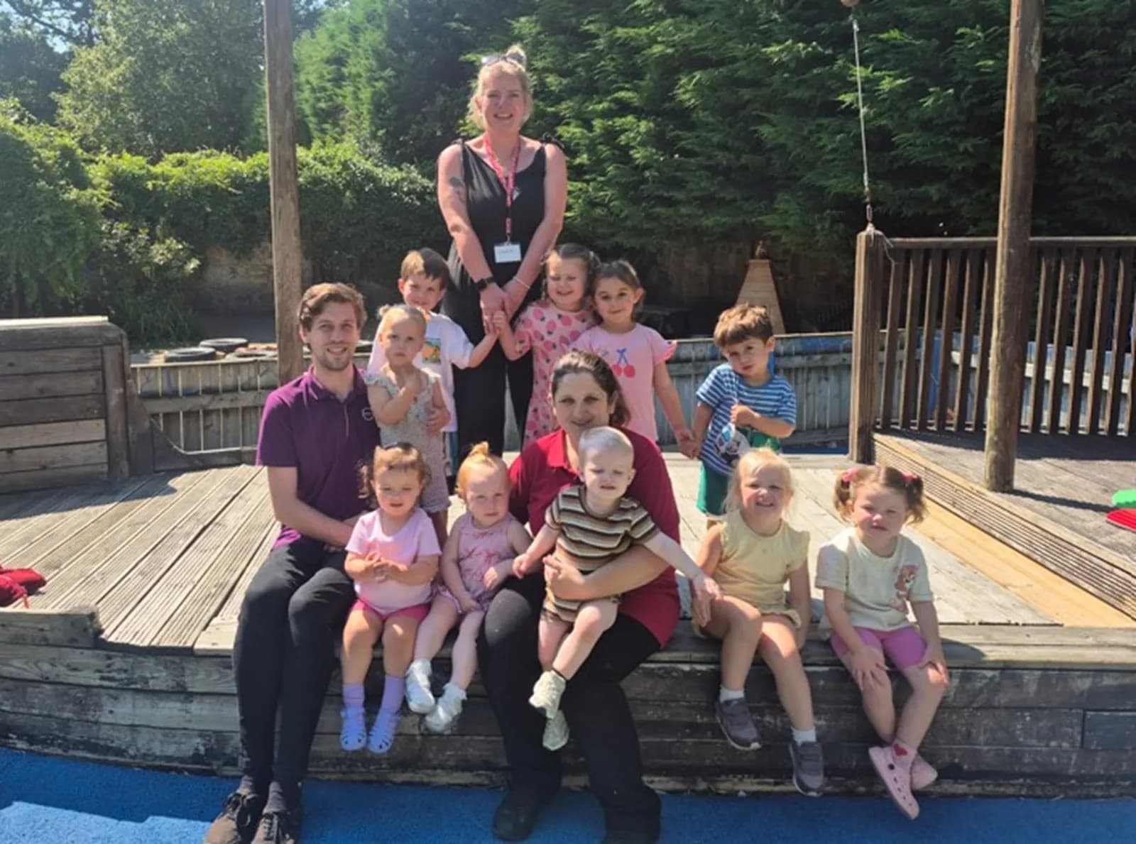 A group of adults and children are gathered in an outdoor play area on sunny day, smiling at the camera.