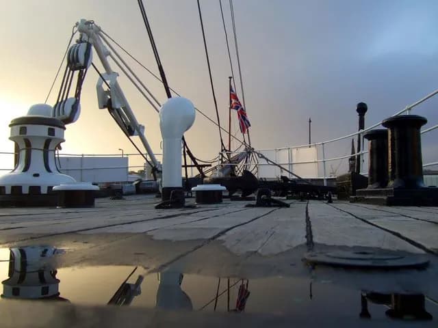 A close-up view of the deck of a historic ship under a cloudy sky, featuring a white winch and crane on the left and a Union Jack flag on the right.