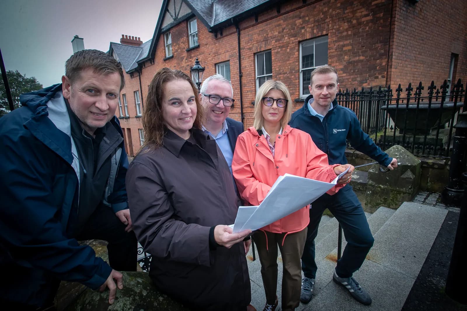 Five adults, three men and two women, stand together outdoors looking at documents, with brick buildings in the background.