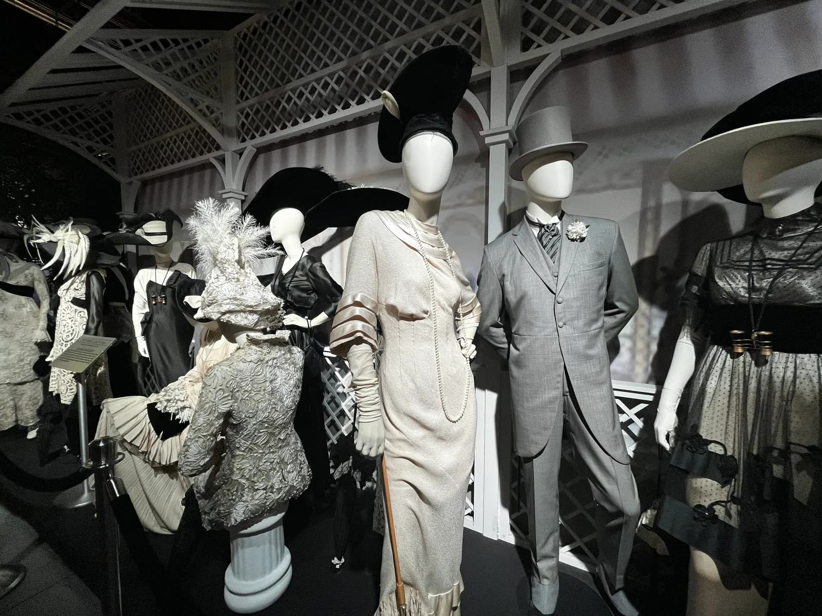 A display of several mannequins dressed in elegant, vintage-style clothing and hats, set up within a white lattice structure.