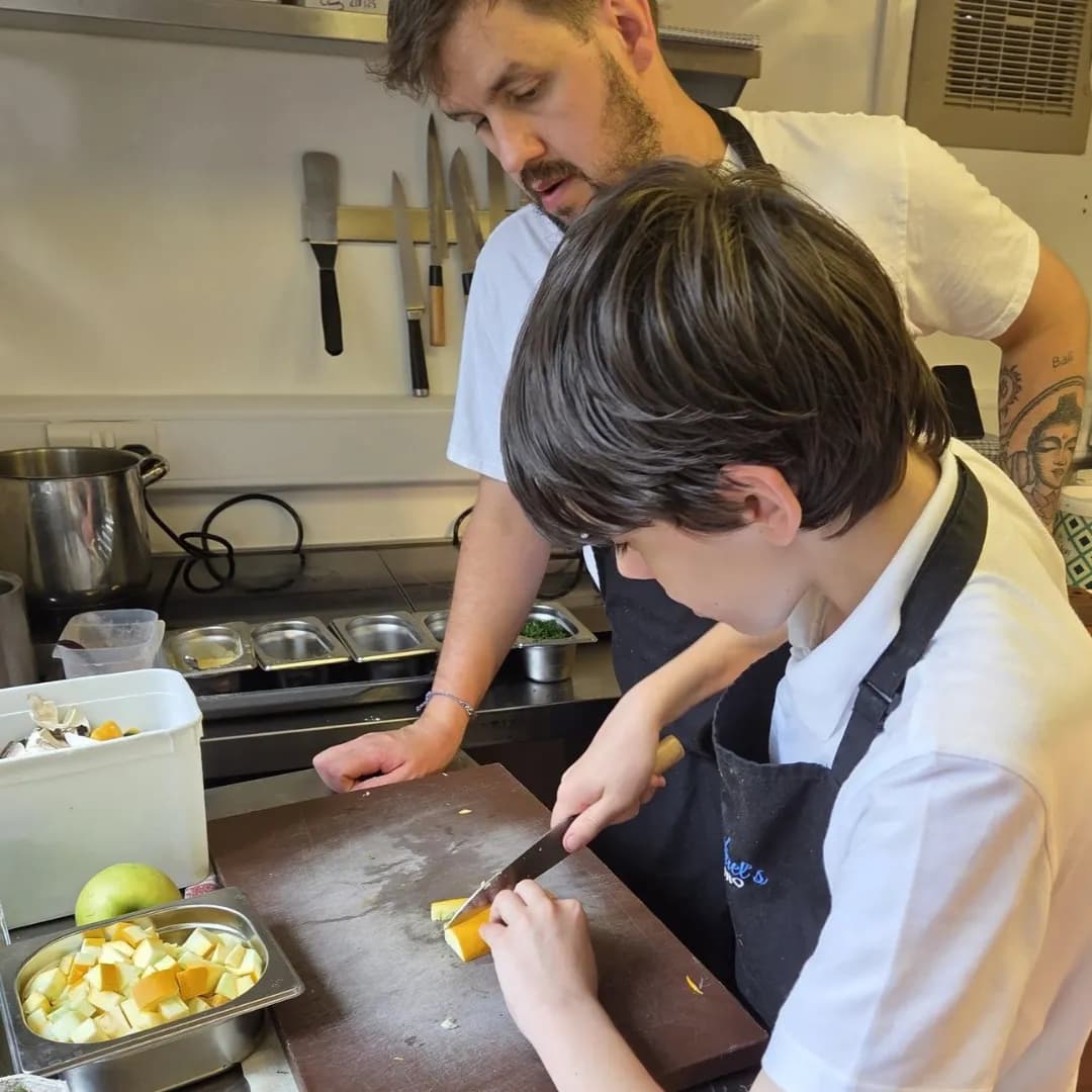 A chef in a black apron and a white shirt instructs a younger person in a black apron and a white shirt on how to chop food on a wooden cutting board in a kitchen.
