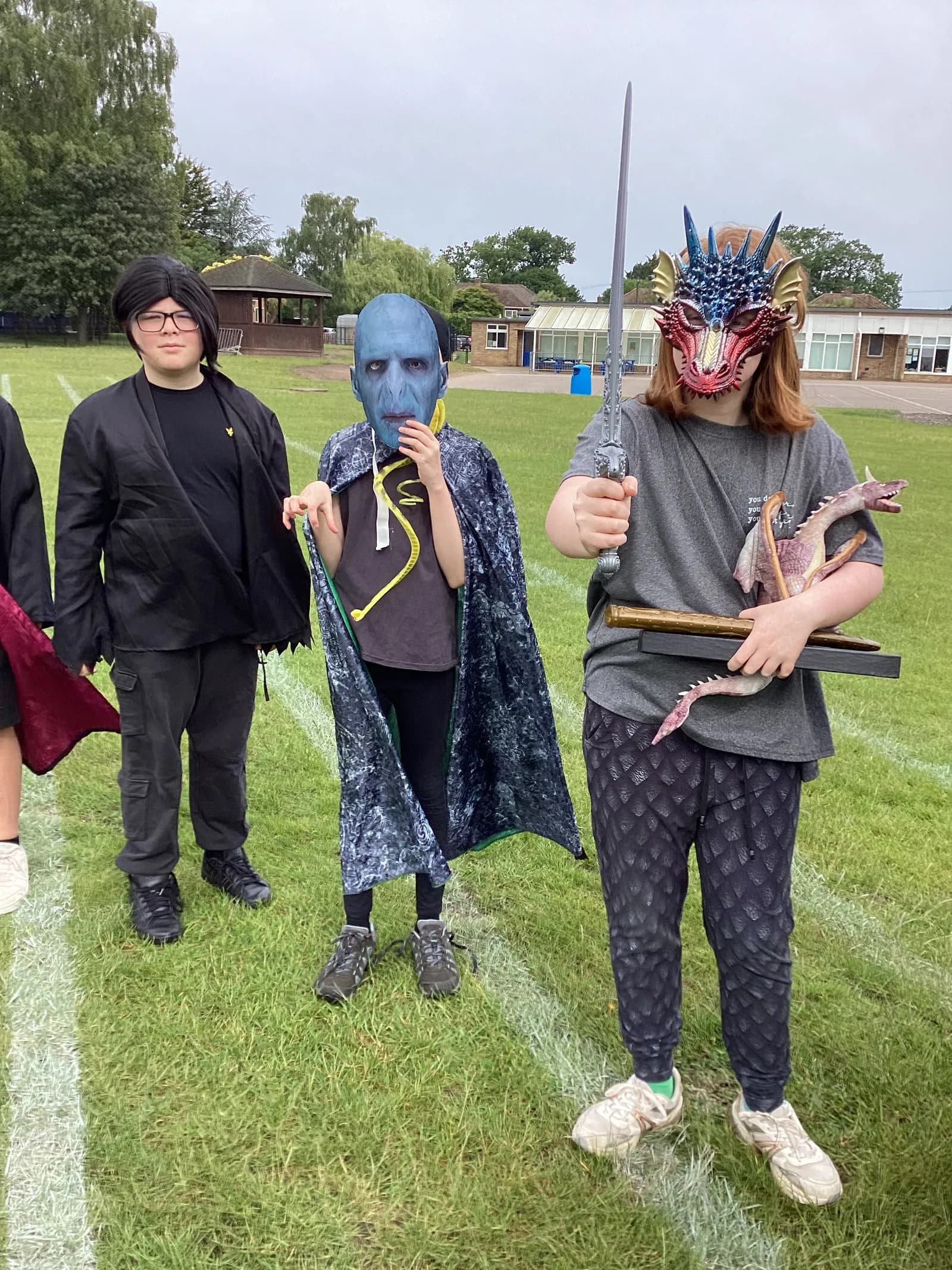 Three children in fantasy costumes, one dressed as Voldemort, stands on a grassy field on a cloudy day.