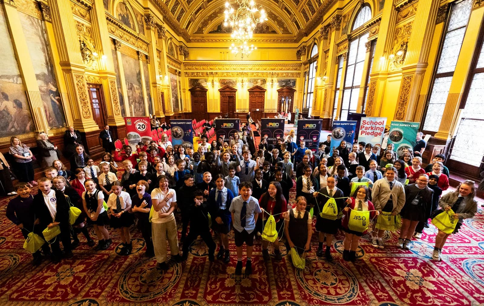 A large group of schoolchildren and adults pose for a picture in a grand hall with ornate gold ceilings and large windows, holding bright yellow bags and signs for 'Road Safety Unit' and 'School Parking Pledge'.