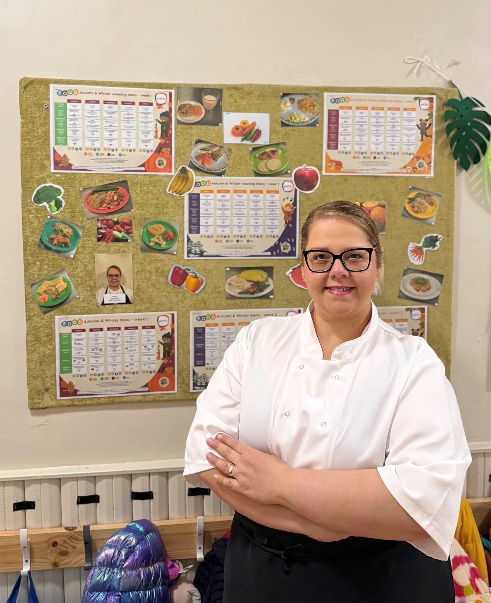 A smiling woman wearing a white chef's jacket and black-rimmed glasses stands with her arms crossed in front of a bulletin board displaying food menus and pictures of dishes.