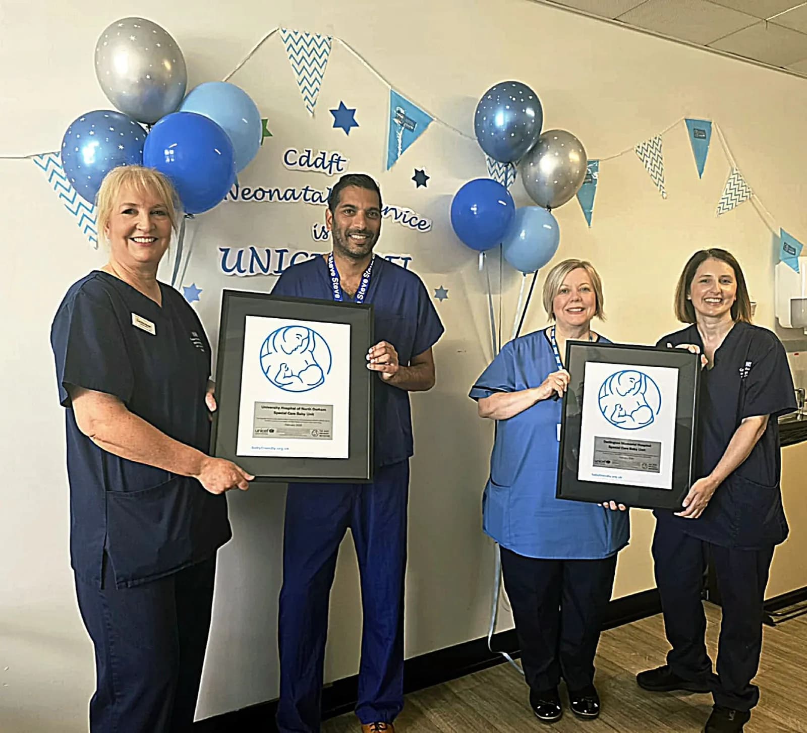 Four healthcare professionals, two men and two women, dressed in blue scrubs, proudly hold up framed certificates indoors, with blue and silver balloons decorating the background.
