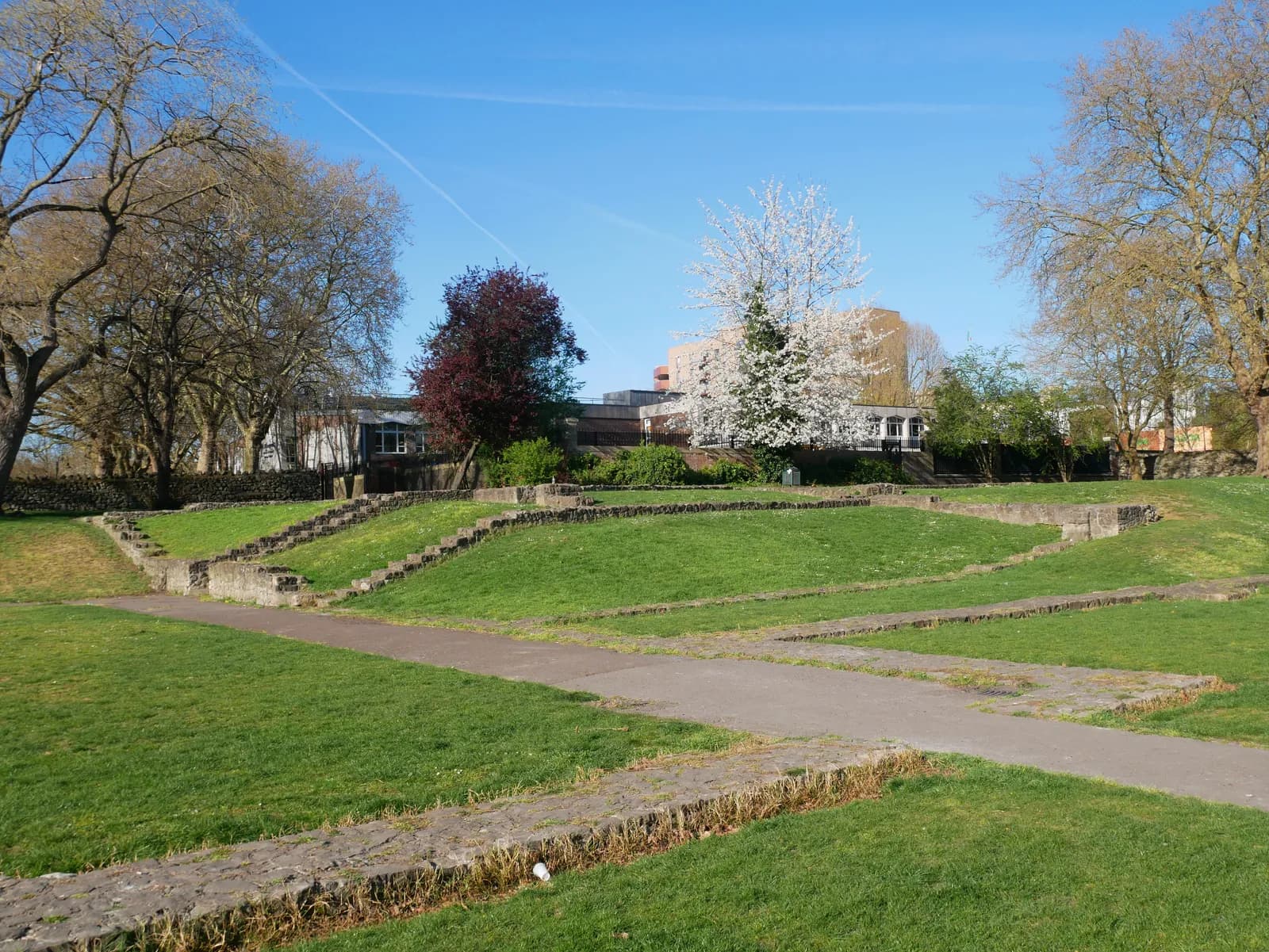 A wide view of a park with green grass and stone pathways leading up to a distant building, framed by trees under a blue sky.